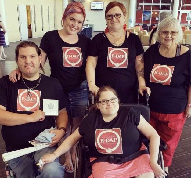 Picture of five Caucasian women with black shirts that have the big red square R-DAP logo on them. Three women are standing and one is kneeling on one knee in front of them and one is sitting in a wheelchair in front of them. They are all smiling.