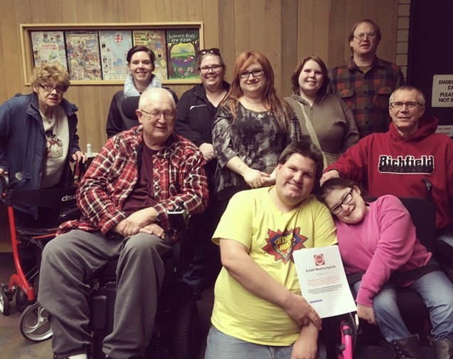 A group photo taken at an R-DAP meeting. This photo is of ten Caucasian men and women, from teenagers to senior citizens. Most are smiling. Two are seated in wheelchairs in front and one woman has a red walker.