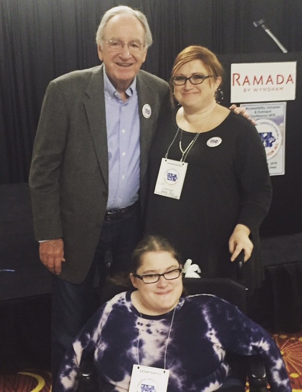 Picture of Former Senator Tom Harkin standing next to Judy Moe, with Raven Moe seated in her wheelchair in front of them. This picture was taken at the first ever Disability Conference for US presidential candidates in Iowa. Former Senator Tom Harkin was the keynote speaker. He was the author of the final version of the Americans with Disabilities Act that was signed into law by President George H. Bush in 1990