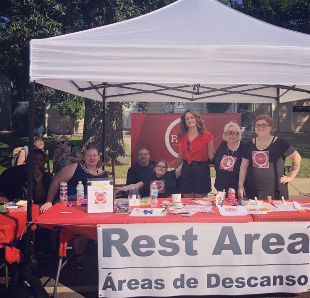 Picture taken at R-DAPS’s booth at Penn Fest 2019. Seven people stand in a line under a white tent, in front of a red banner with R-DAP’s logo on it, behind a red table with a sign hanging that says “Rest Area” and “Areas de Descanso” on it. The seven people from left to right: Daquan, Kaylyn, Kevin, R-DAP co-founder Raven Moe, Richfield Mayor Maria Regan Gonzalez, Evie, and R-DAP co-founder Judy Moe.