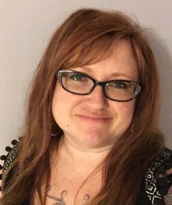 Picture is a headshot of Judy Moe, the Director, and Co-founder of Richfield Disability Advocacy Partnership. She has long auburn hair, black framed glasses, with rhinestones on the outside edge of frames. She is wearing a black, sleeveless top. She is smiling.